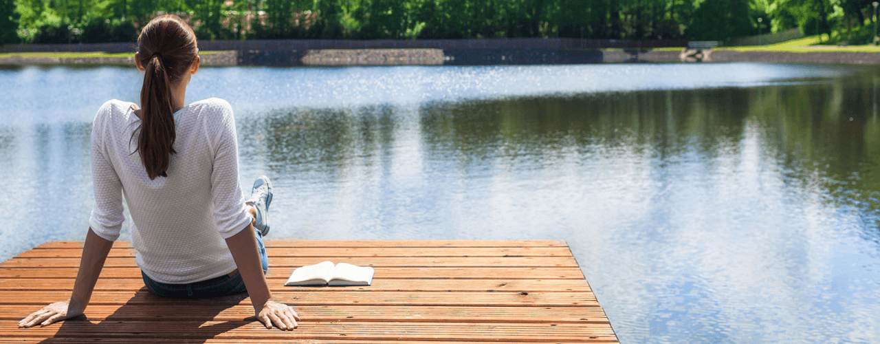 A back view of a girl sitting on a dock looking out at the calm water.