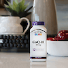 A bottle of 21st Century Co Q-10 100 mg capsules sitting on a kitchen counter with a bowl of apples, potted plant and vase in the background.