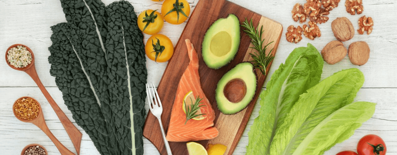 Top view of brain healthy foods like avocado, kale, tomatoes on a white wood background.