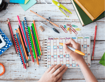 Top view of back-to-school items like pencils, a calendar, paper clips, books all on a white wood background.