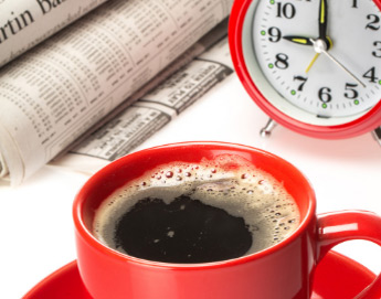 A red coffee cup filled with coffee next to an analog clock and newspaper.