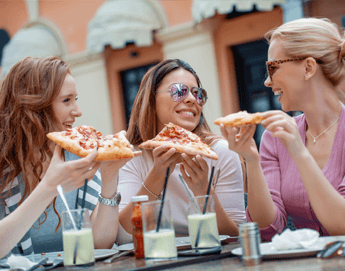 A group of friends seated at an outdoor patio laughing and eating pizza.