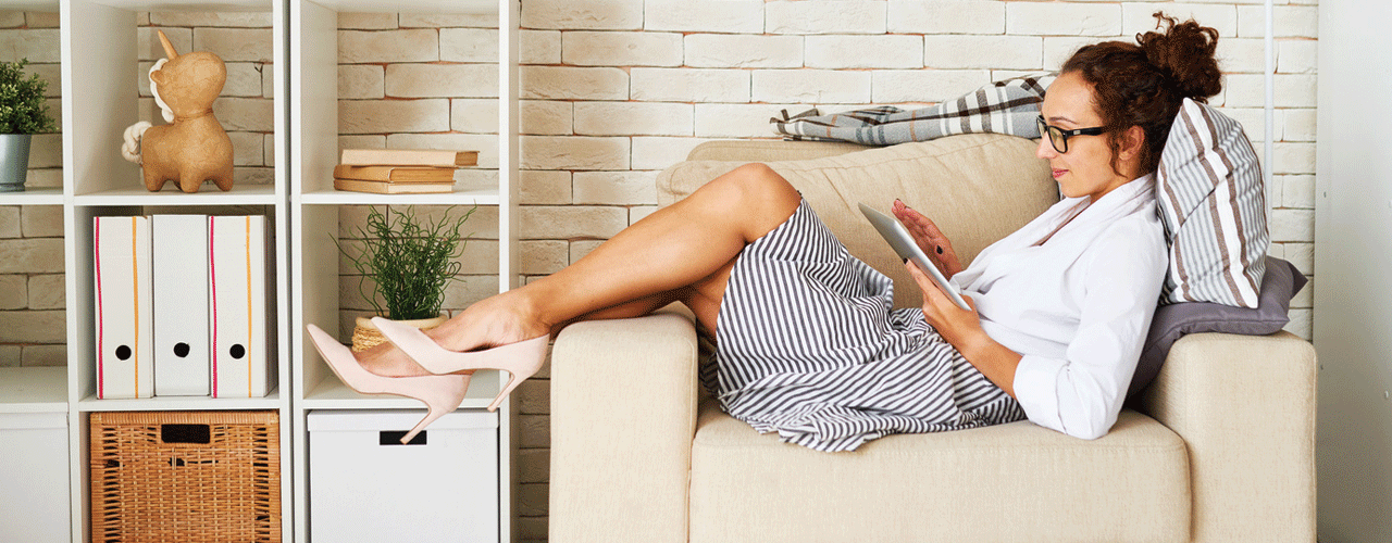 A girl laying across a living room chair with her legs up on the chair arm while looking at an iPad.