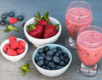 Two glasses filled with smoothies next to bowls of strawberries, blueberries and raspberries.