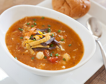 A bowl tortilla soup with spoon and bread roll.