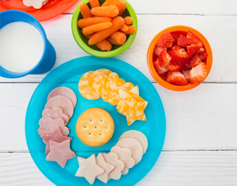 A top view of a plate with lunch meat cutouts, carrots in a bowl, and strawberries in bowl.