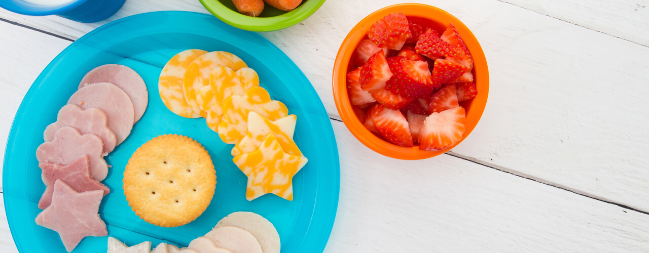 A top view of a plate with lunch meat cutouts, carrots in a bowl, and strawberries in bowl.