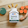 A bottle of 21st Century Turmeric 500 mg Complex vegetarian capsules laying on a wood background with capsules coming our of the top and turmeric root to the right.